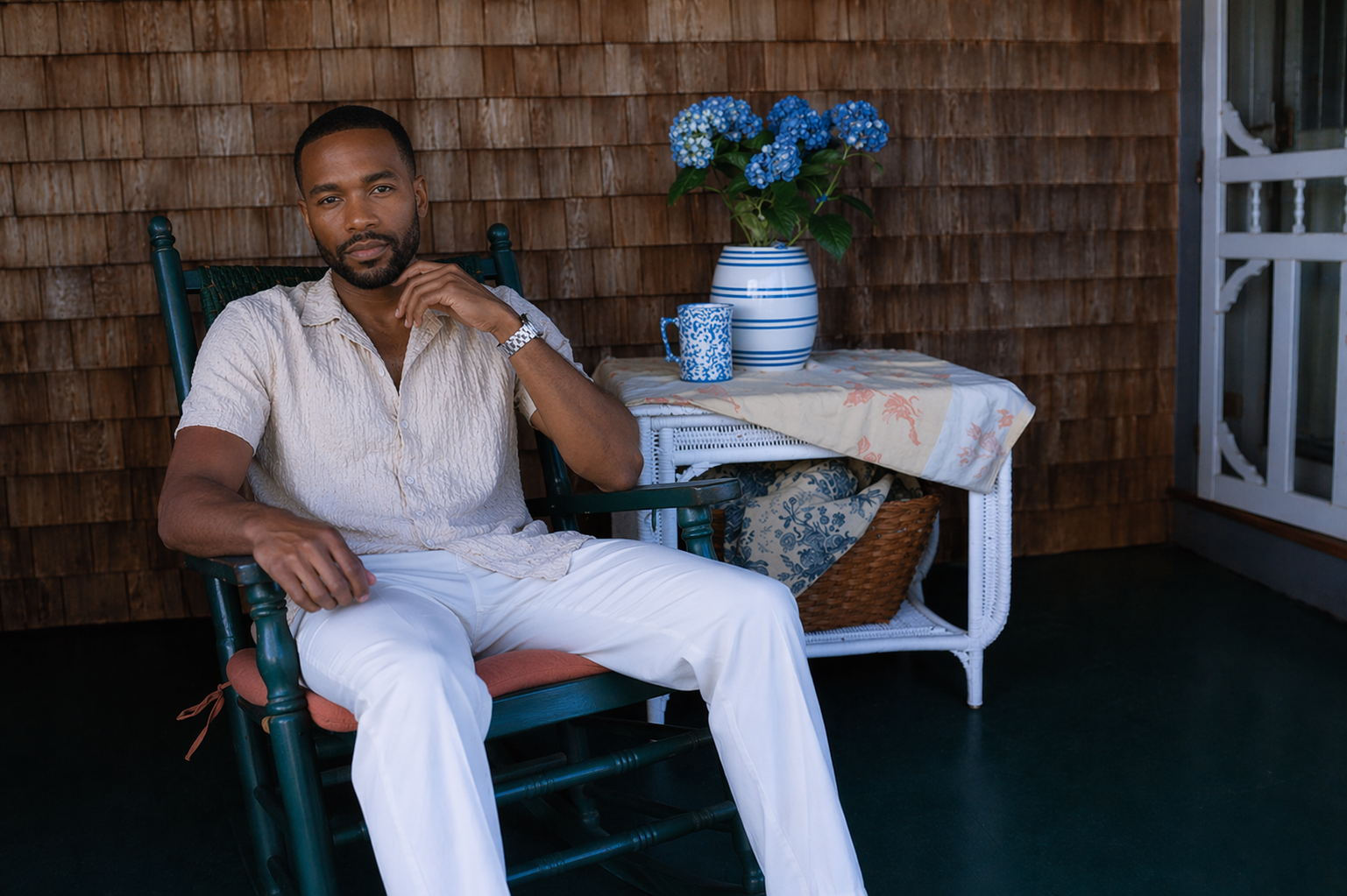 Malik Carter, founder of Black Vineyard Events, seated on a Martha's Vineyard porch rocking chair in summer style with cedar shingles, hydrangeas, and coastal New England porch details.